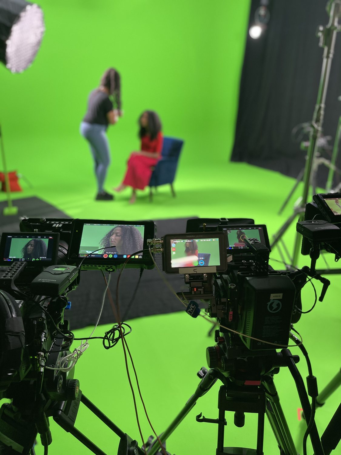 Studio cameras and their monitors framing talent being prepped on a green-screen set