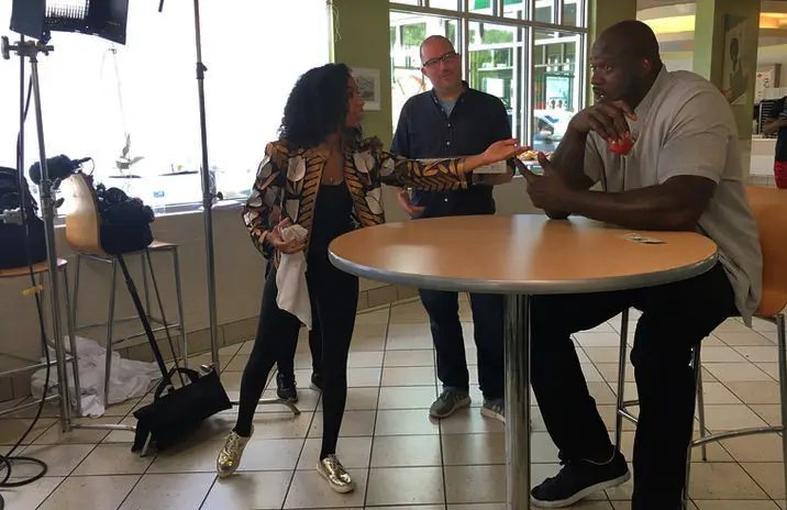 Chelsea coaching Shaquille O'Neal at a table on the set of a Dunkin' commercial
